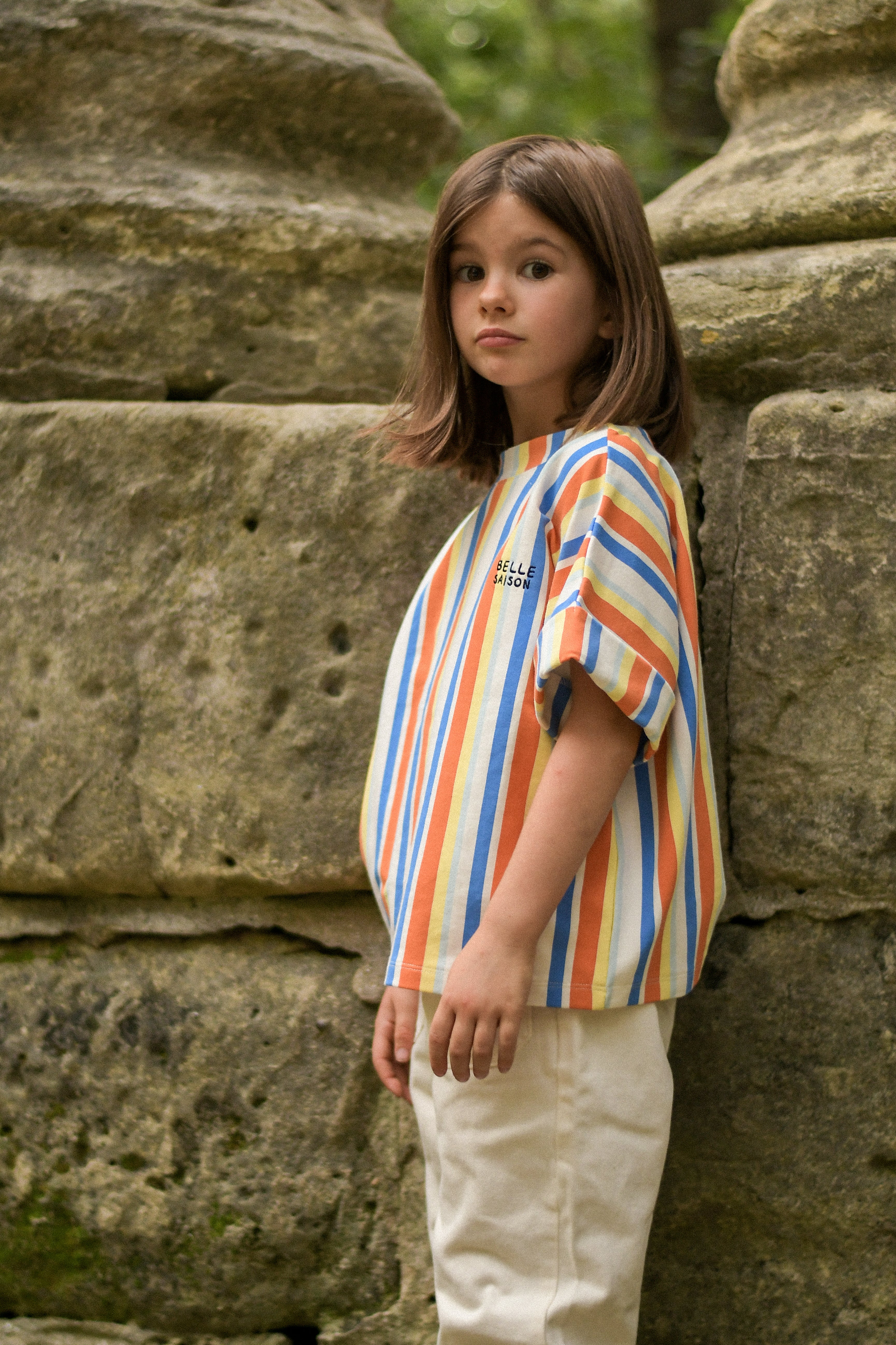 Model wearing striped boxy style t-shirt in multiple colours (red, blue, yellow, orange, and white). The t-shirt has a round neckline and the phrase "Belle Saison" on the left side. Made by Maison Tadaboum